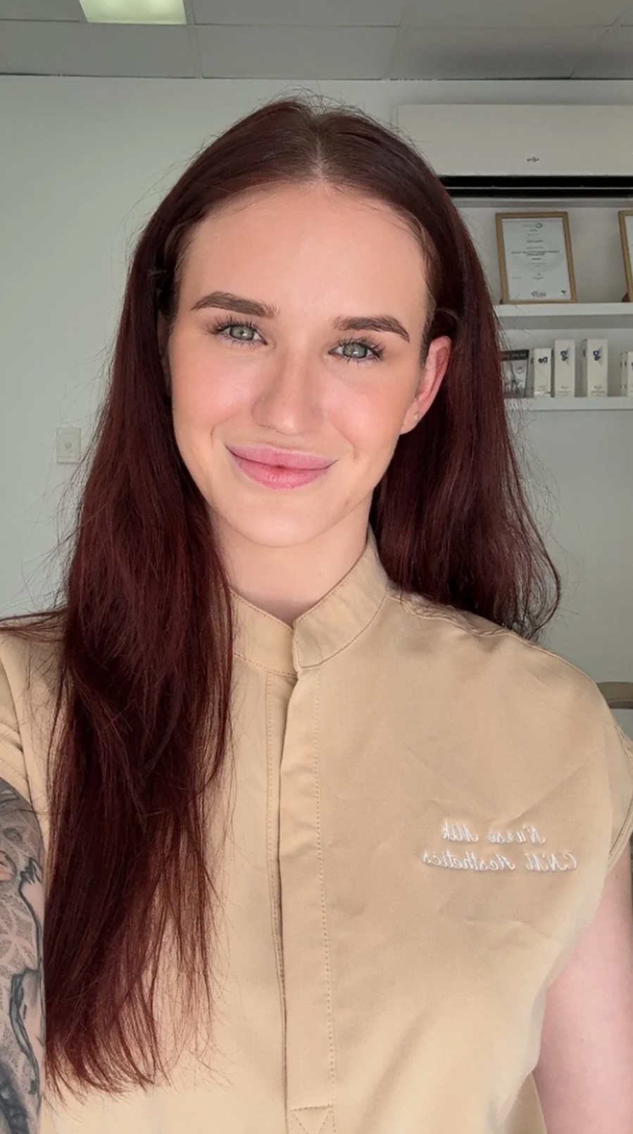 A smiling woman with long red hair, light skin, and blue eyes, wearing a beige uniform with white embroidered text on the chest, in an indoor setting with certificates and white shelves in the background.
