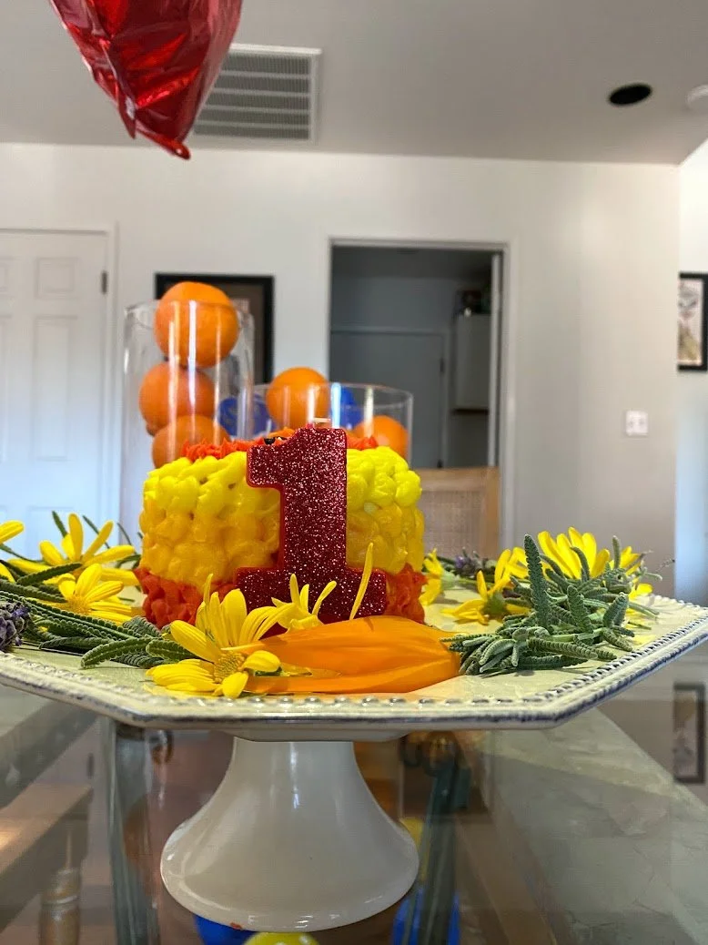 A birthday cake decorated with yellow, red, and purple icing, topped with a red glitter number one candle. The cake is on a decorative white cake stand and surrounded by yellow flowers and green foliage. In the background, there are tall clear vases