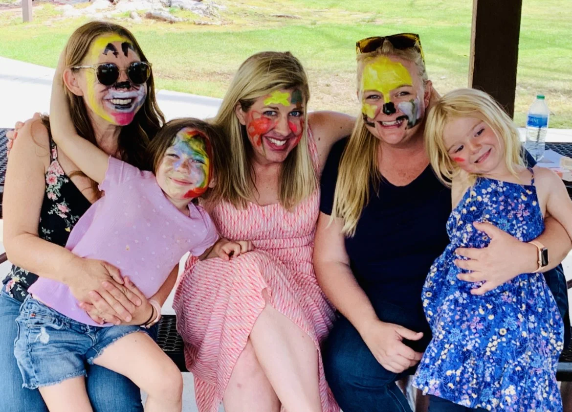 Group of five women and two young girls with faces painted in bright colors, smiling and enjoying each other's company outdoors under a pavilion, with a grassy area in the background.
