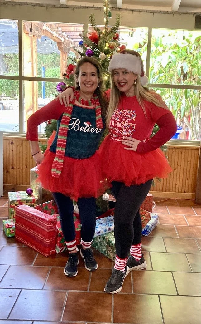 Two women dressed in festive holiday outfits, wearing red tutus, standing in front of a decorated Christmas tree. They are indoors with large windows, surrounded by Christmas presents. One woman is wearing a Santa hat, and both are smiling.