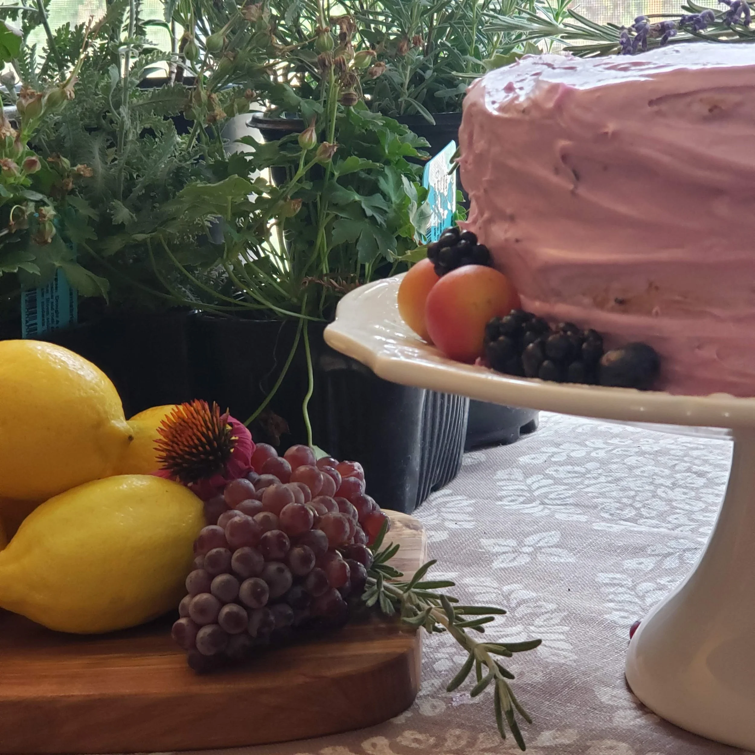 A table decorated with a pink frosted cake topped with small fruits including blackberries and apricots, next to a wooden platter holding lemons, grapes, a flower, and sprigs of herbs, with green plants in the background.