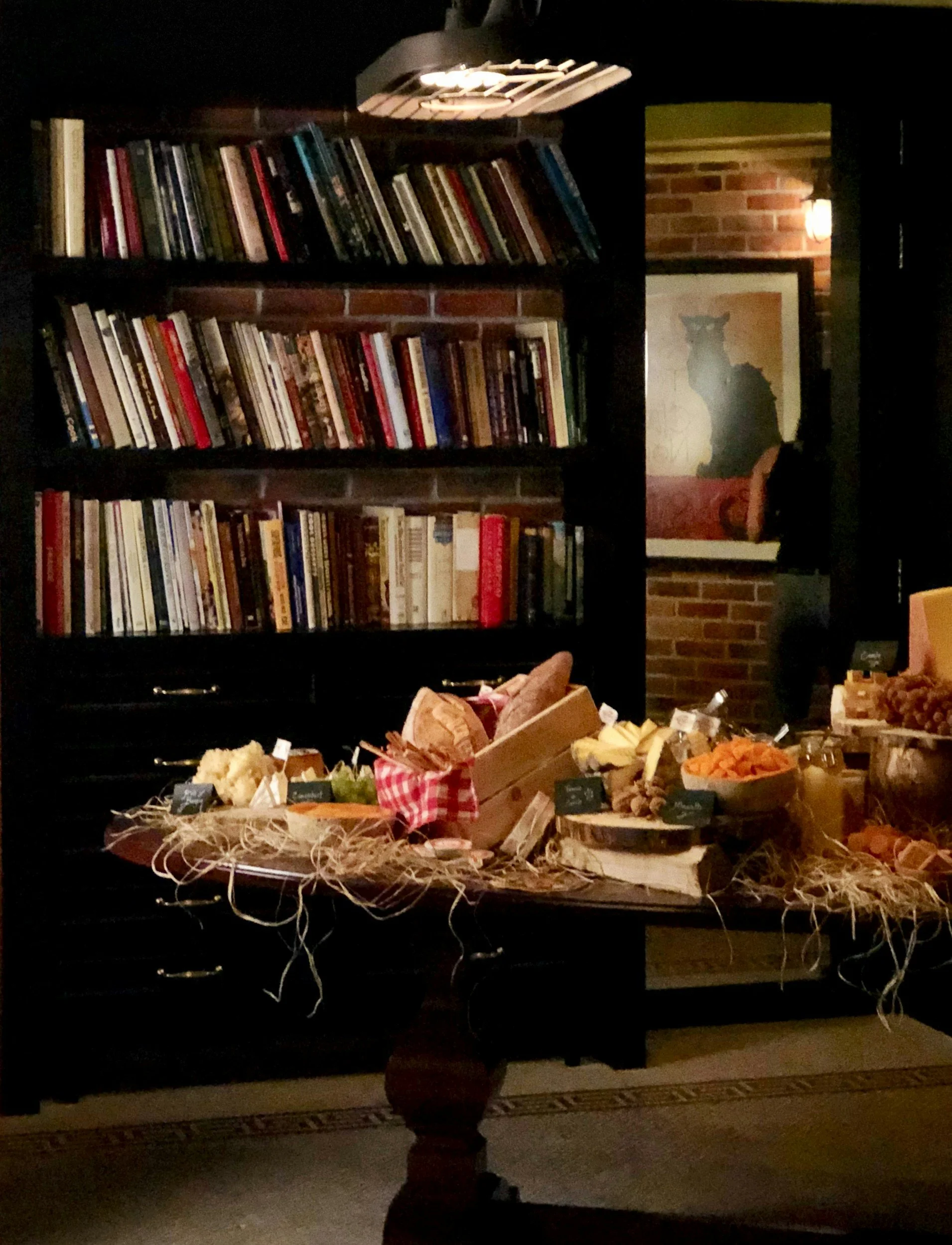 A dark interior scene with a wooden bookshelf filled with books, a framed cat picture on brick wall, and a table with assorted food items, produce, and small signs.