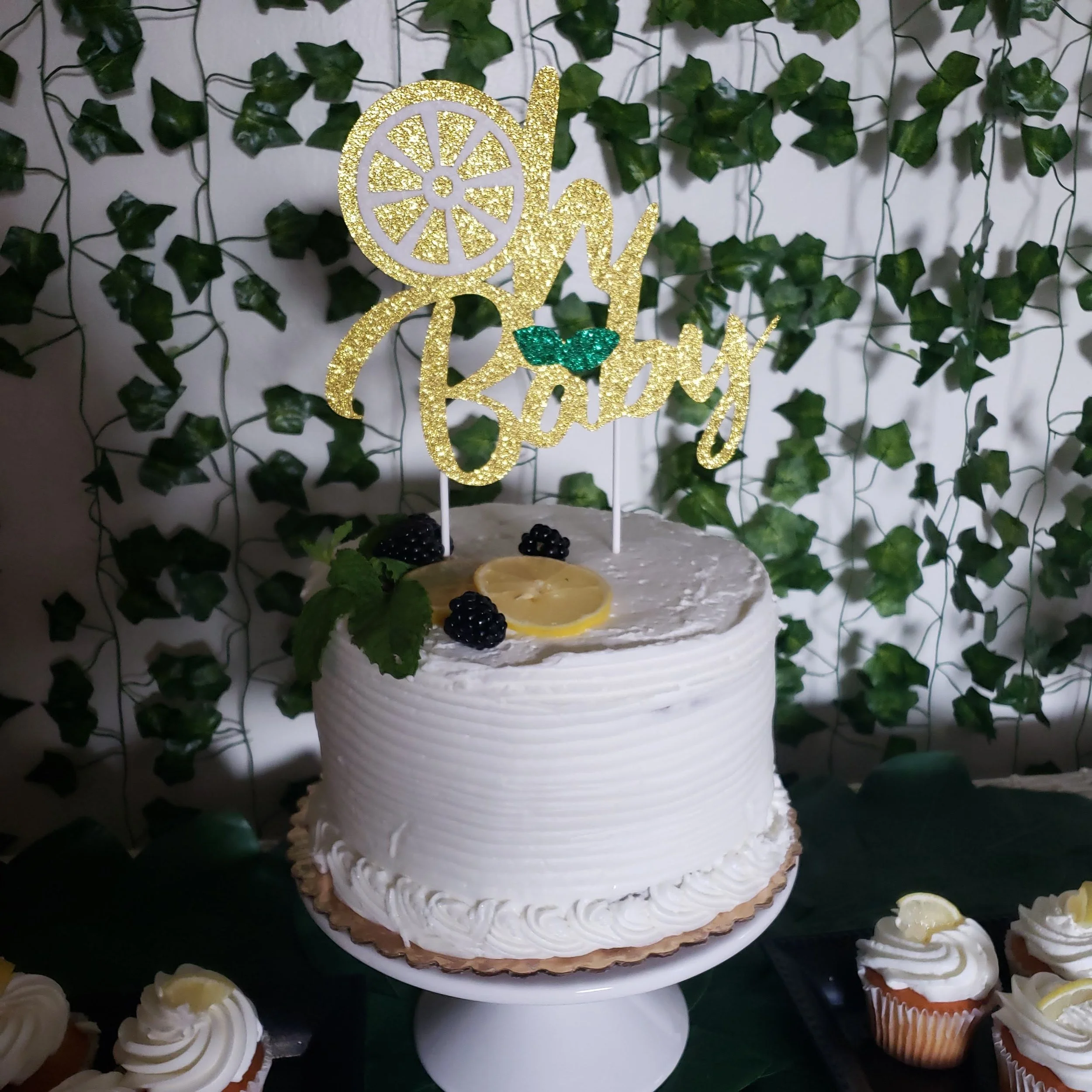 White frosted birthday cake with lemon slices and blackberries, topped with a glittery gold "Happy Birthday" topper, against a green ivy background.
