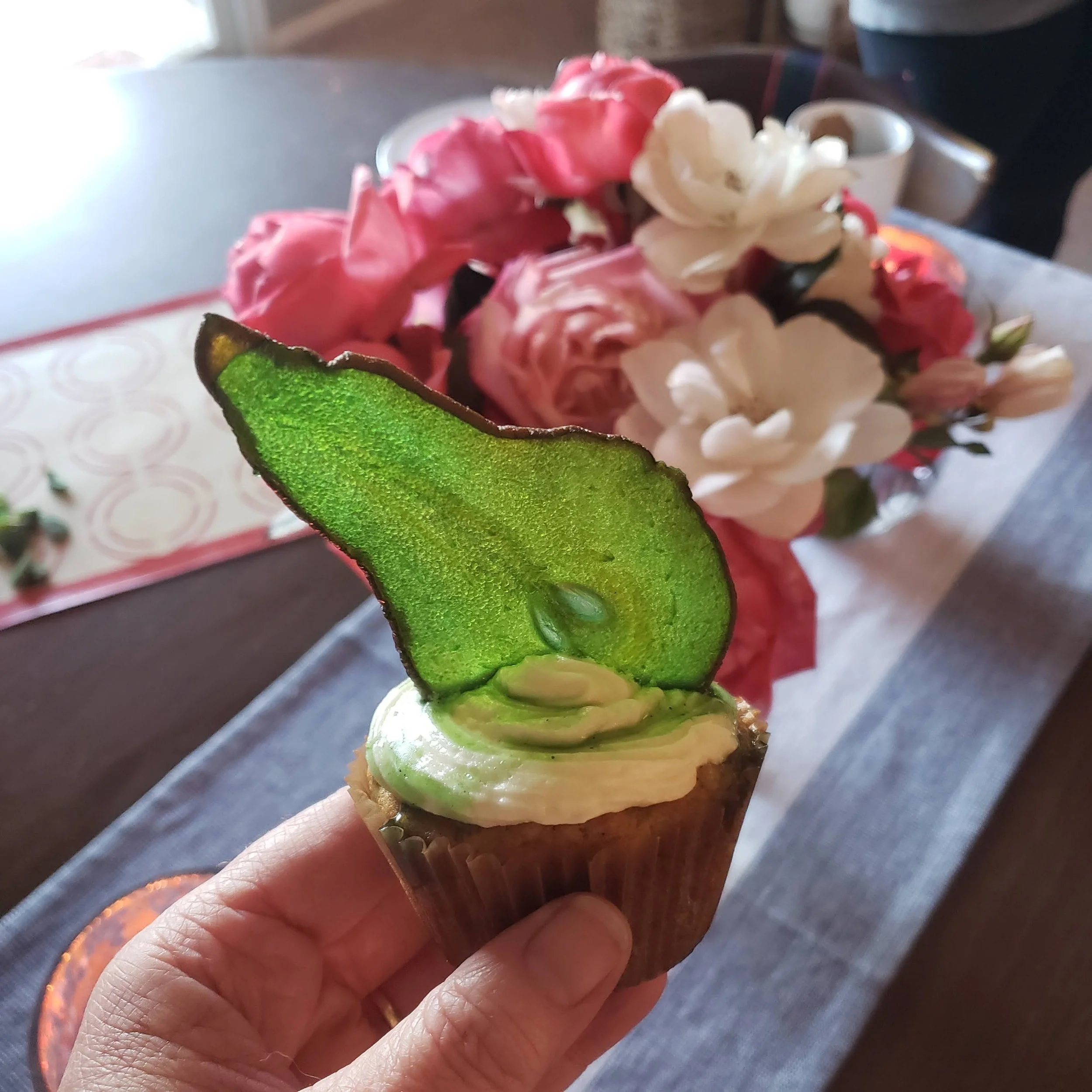 A cupcake with green frosting and a slice of fresh green apple on top, held by a hand in front of a flower arrangement on a table.