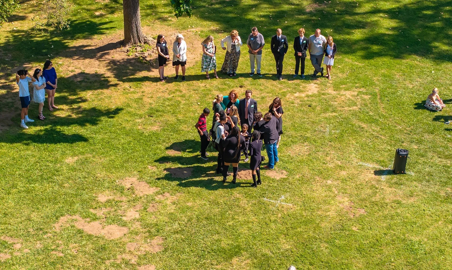 A group of people gathering outdoors on a grassy area, some standing and some sitting, in what appears to be a wedding ceremony. There is a large tree and a speaker visible in the scene.