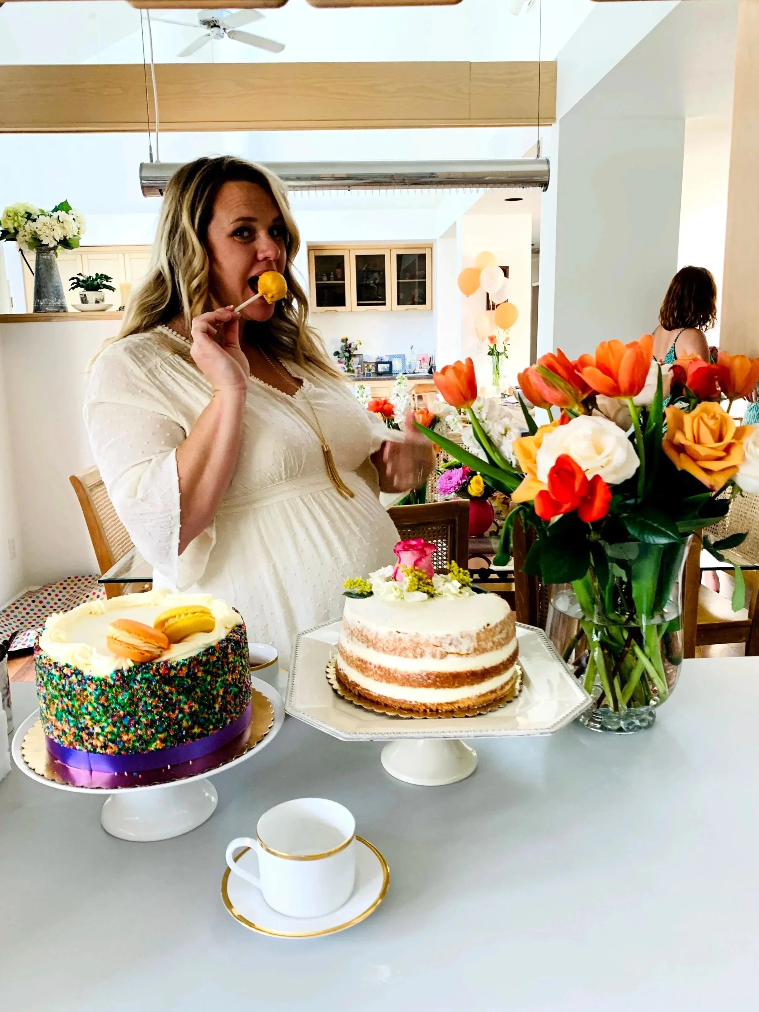Woman in a white dress at a celebration table with cakes, flowers, and a teacup, holding a yellow cake pop near her mouth.