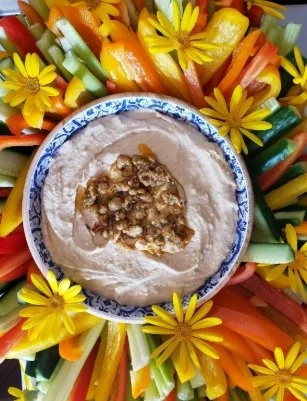 A bowl of hummus with toppings, surrounded by sliced carrots, cucumbers, bell peppers, and yellow flowers.