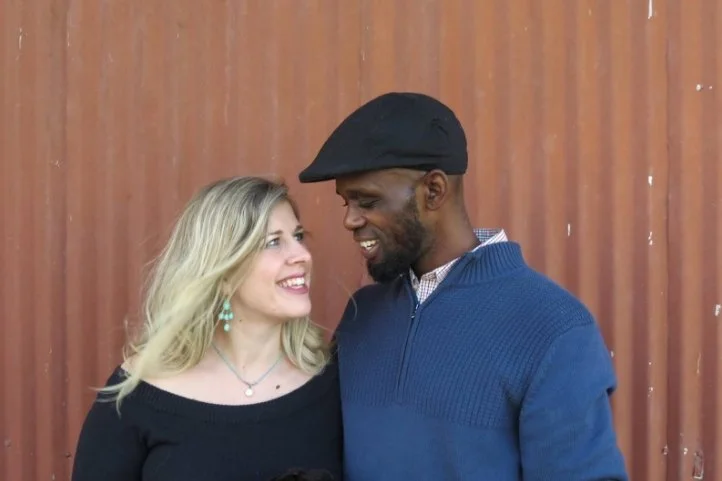 A woman and a man smiling at each other outdoors with a wooden fence background.