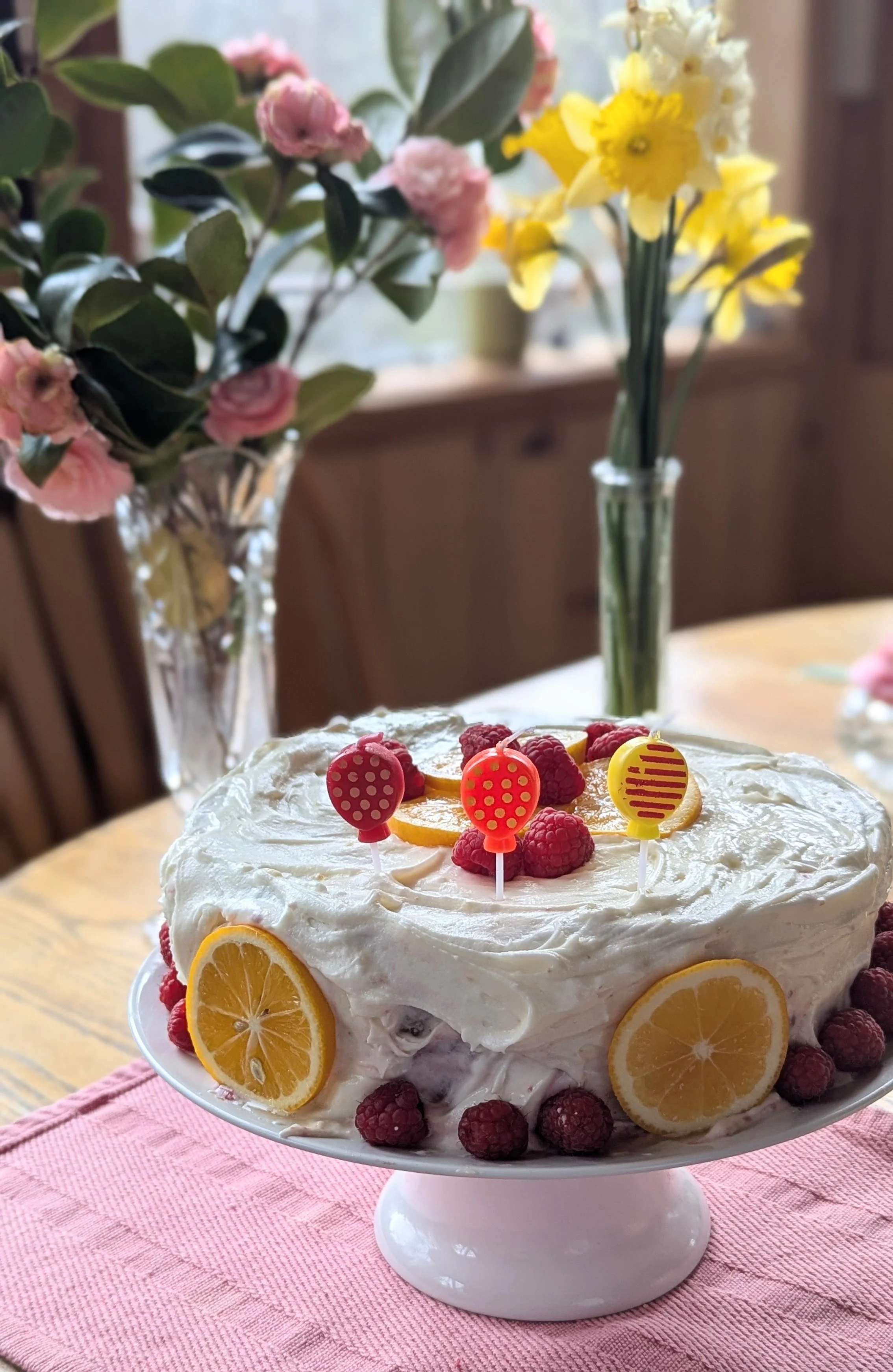 A decorated lemon cake with raspberries and lemon slices, topped with colorful candle-holders, on a white cake stand on a pink cloth, with pink and white flowers in vases in the background.