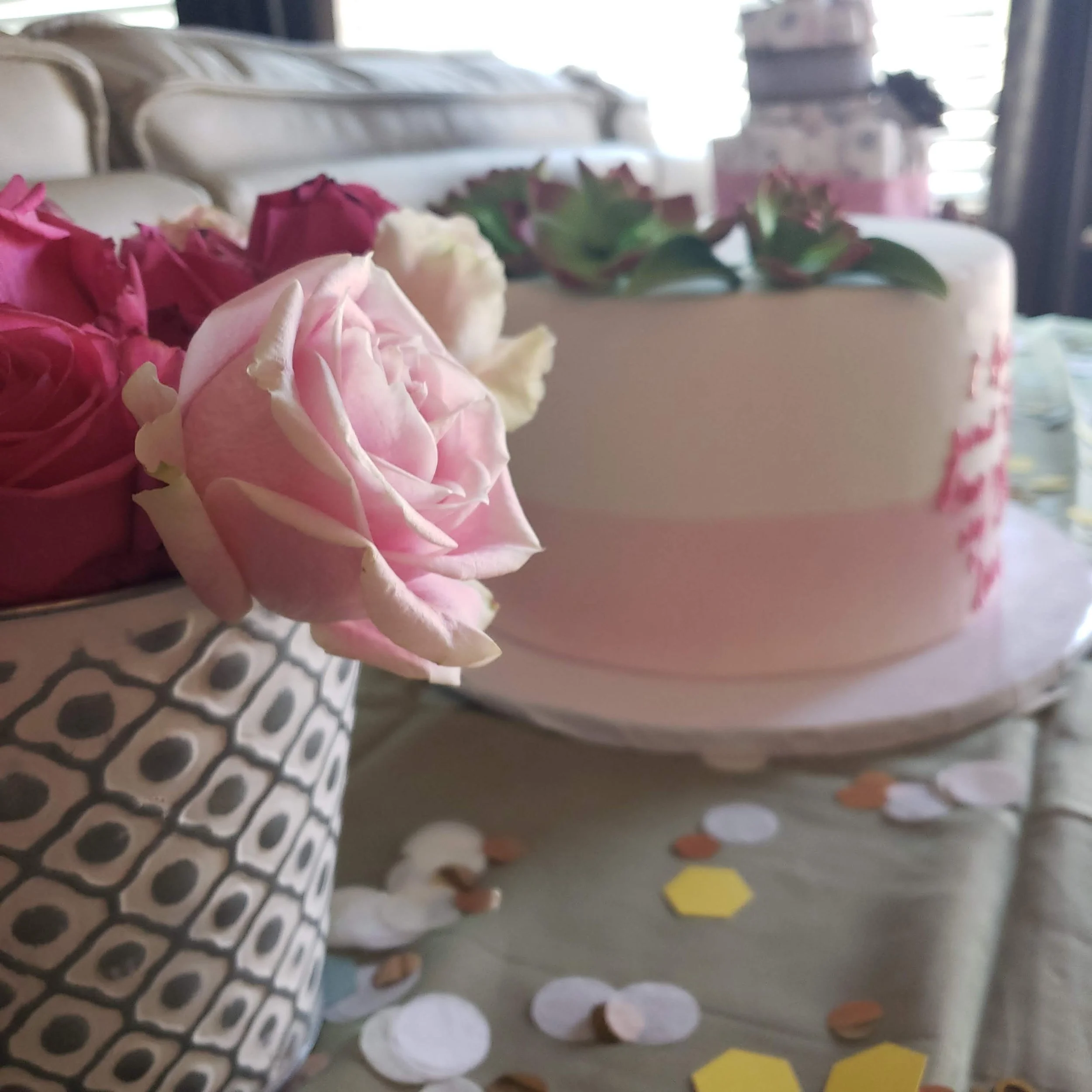 Close-up of pink and white roses in a patterned vase in front of a decorated cake with pink and white floral accents, on a table with scattered confetti.
