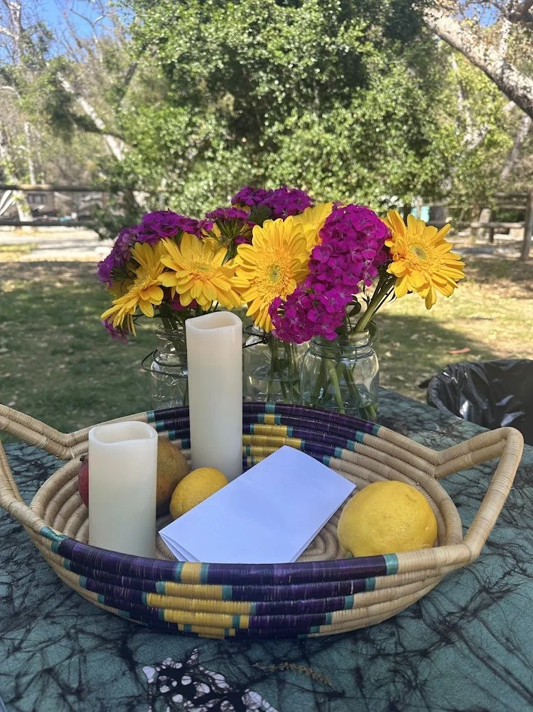 A table outdoors with a woven basket containing two cloves of lemon, an apple, and a stack of white paper, with two white candles beside the basket. Behind the basket, there are glass jars filled with yellow and purple flowers. The background shows t
