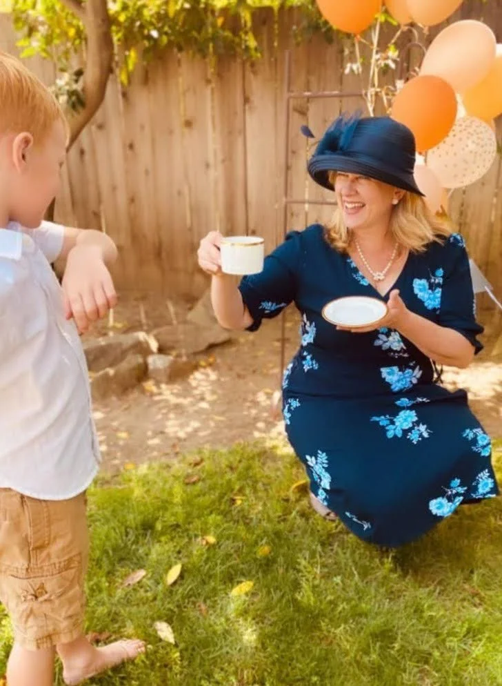 A woman in a blue floral dress and black hat is kneeling on the grass, smiling and holding a teacup and saucer. A young boy with red hair wearing a white shirt and tan shorts is standing nearby. There are balloons and a wooden fence and plants in the background, suggesting a garden or outdoor party setting.