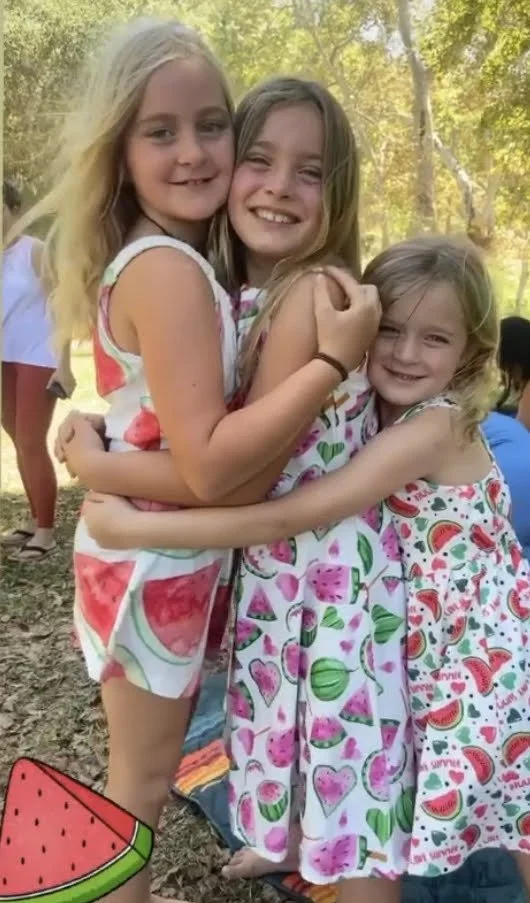 Three young girls hugging outdoors during daytime, dressed in colorful summer dresses with fruit and watermelon patterns, surrounded by trees and other children in the background.