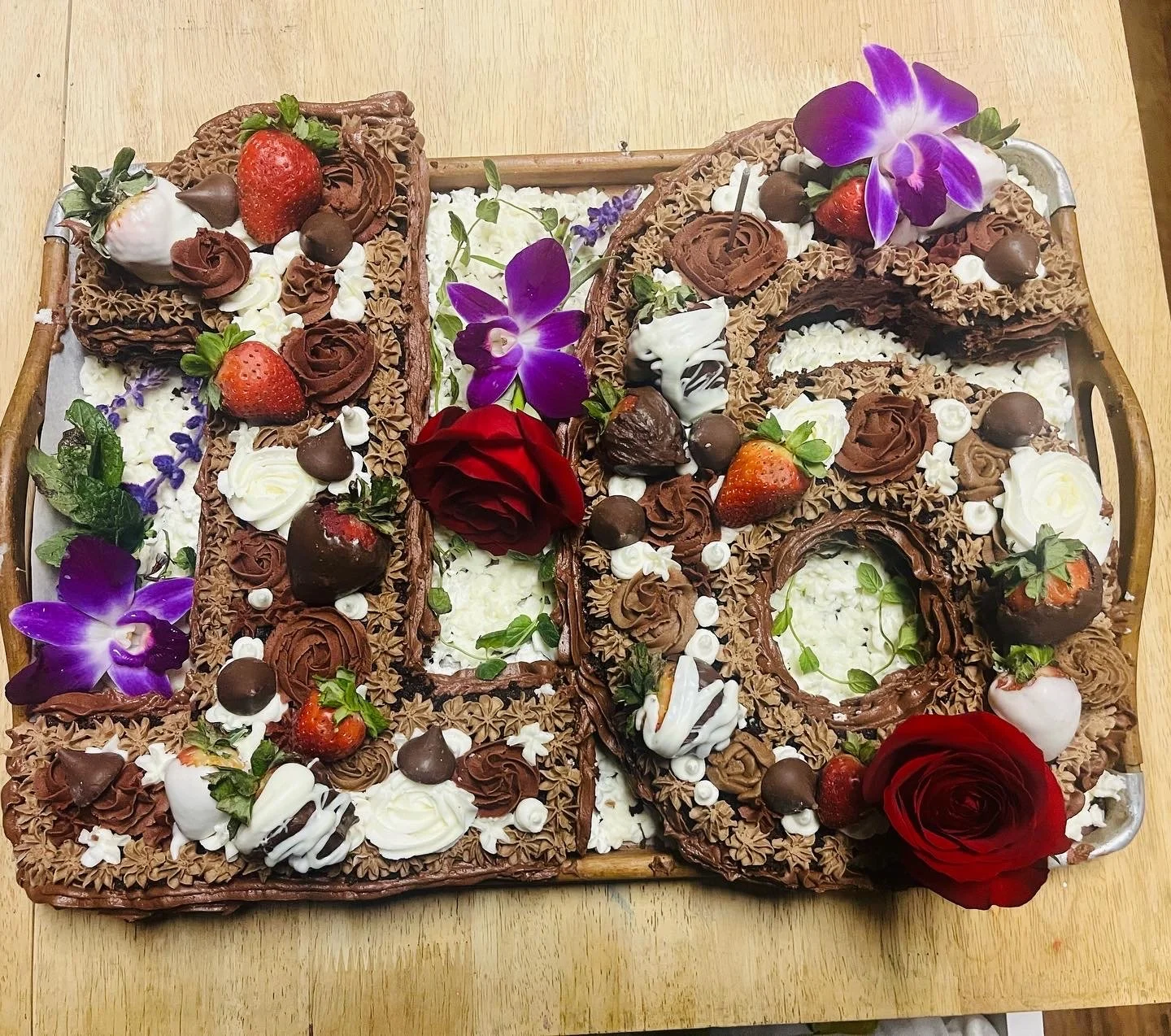 Large cake resembling a large number five decorated with strawberries, chocolate rosettes, flowers, and roses, on a wooden table.
