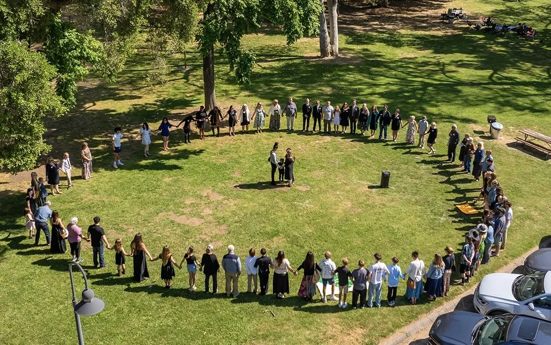 A large group of people is standing in a circle on a grassy area, possibly for a wedding or community gathering, with a couple at the center exchanging vows or rings, surrounded by trees and parked cars.