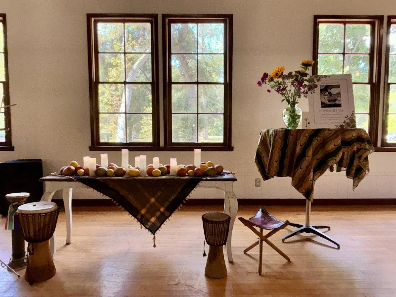 Room with three large windows, a table with candles and fruit, a flower arrangement, and a music stand with a framed picture and cloth, all with natural light.