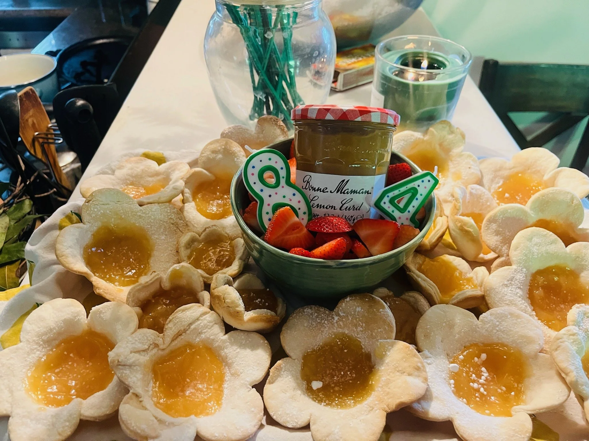 A table with flower-shaped cookies filled with yellow jam, a jar of lemon curd, and strawberries in a green bowl. There is also a clear vase with green pencils, a candle, and some cups in the background.
