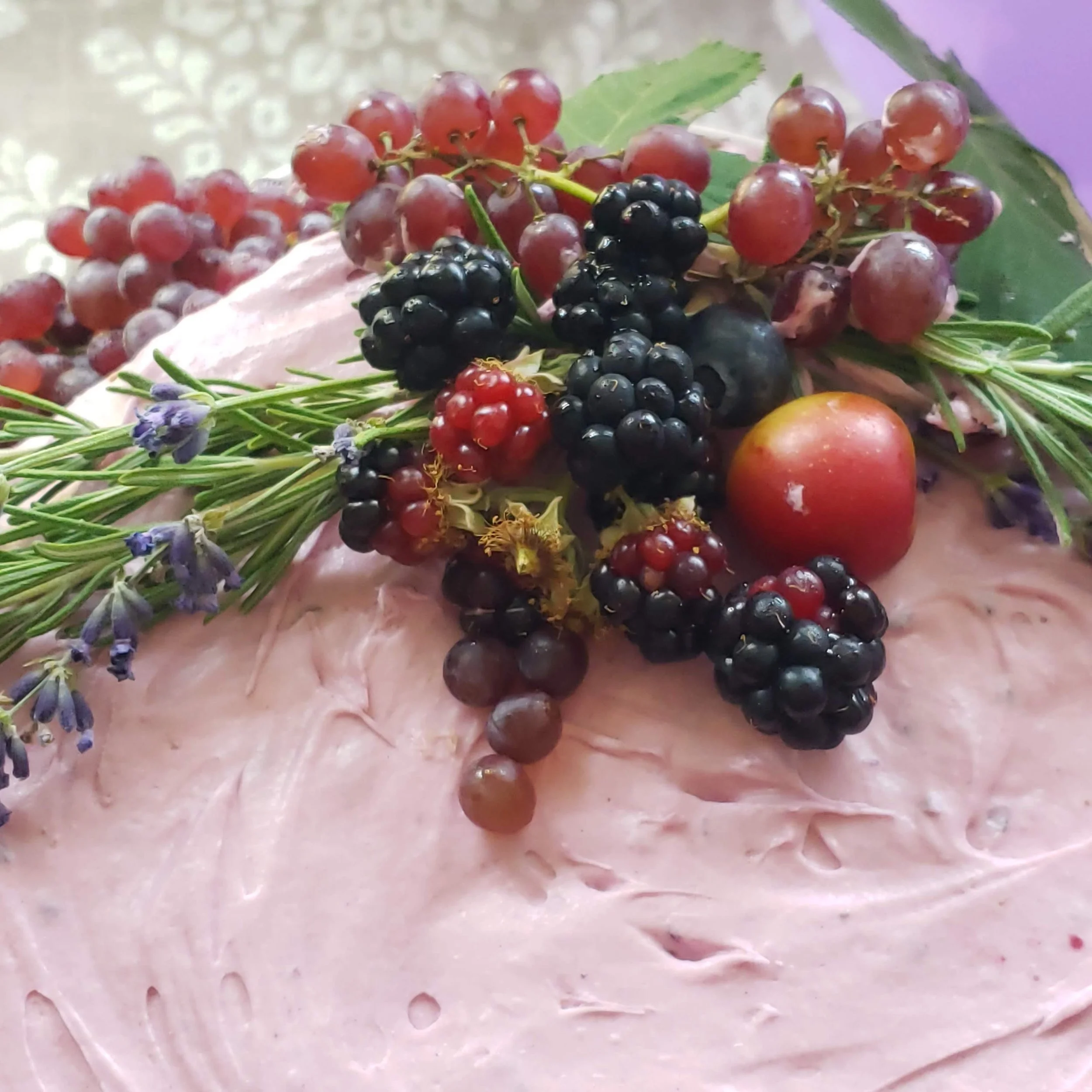 Frosted cake decorated with blackberries, red currants, grapes, an apple, lavender sprigs, and green leaves.