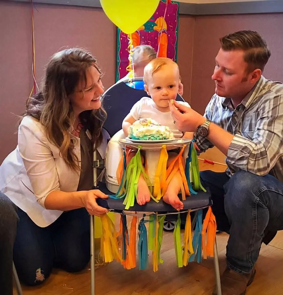 A birthday celebration with a toddler in a high chair decorated with colorful streamers, with a woman and a man feeding the child cake, while balloons and birthday decorations are in the background.