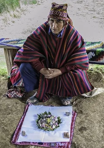 Peruvian Shaman wearing a colorful striped poncho, on the ground with hands clasped, next to a small ceremony altar with flowers and candles.
