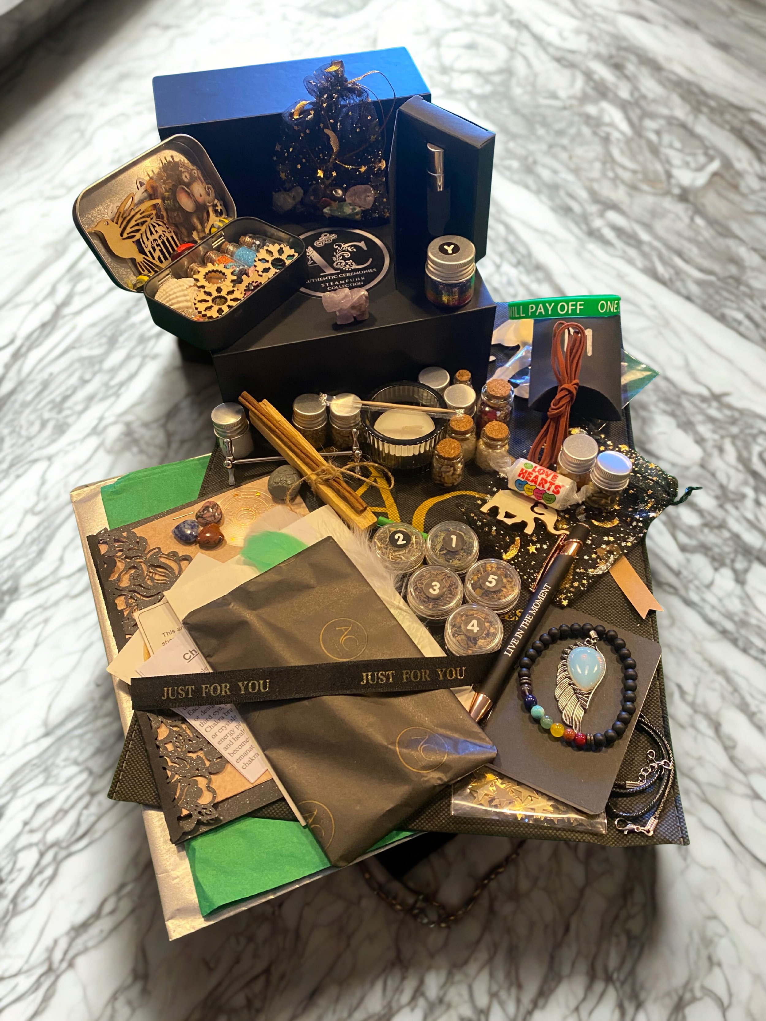 Collection of holistic and spiritual items including essential oils, crystals, herbal bundles, wooden and metal charms, candles, and a decorative box, arranged on a marble surface.
