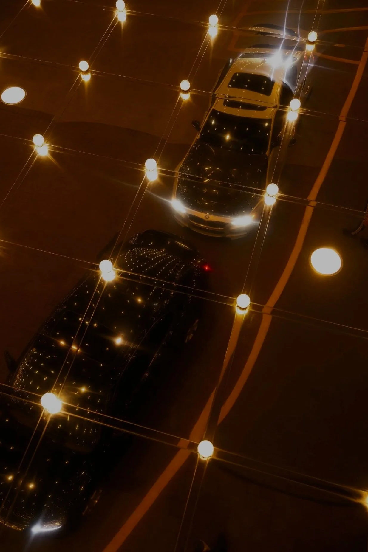 Overhead view of two black cars parked in a parking lot at night, with string lights creating starburst reflections on the cars.