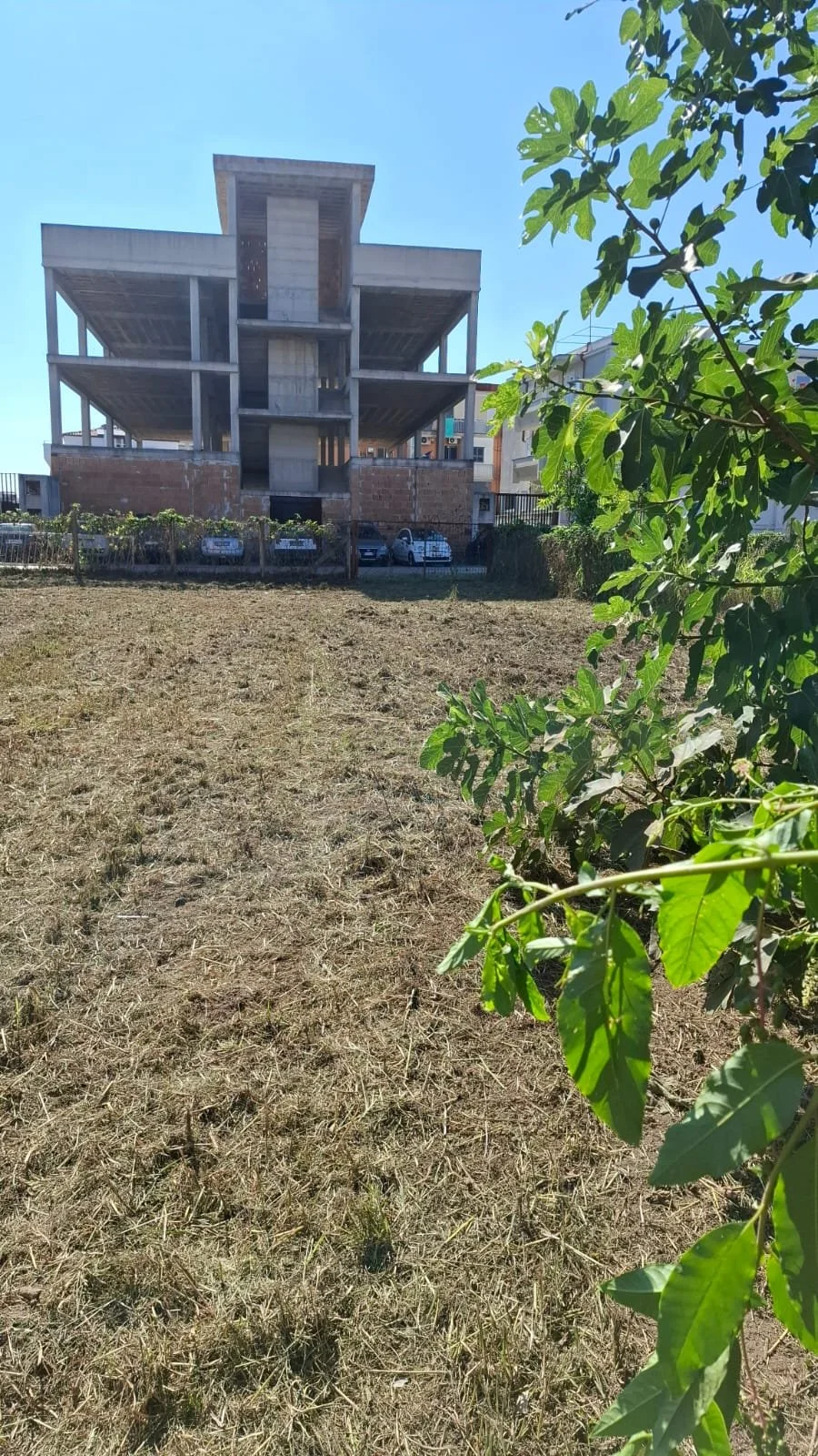 Costruzione di un edificio incompleto con un'area di terreno vuoto davanti e alcune piante verdi sulla destra.