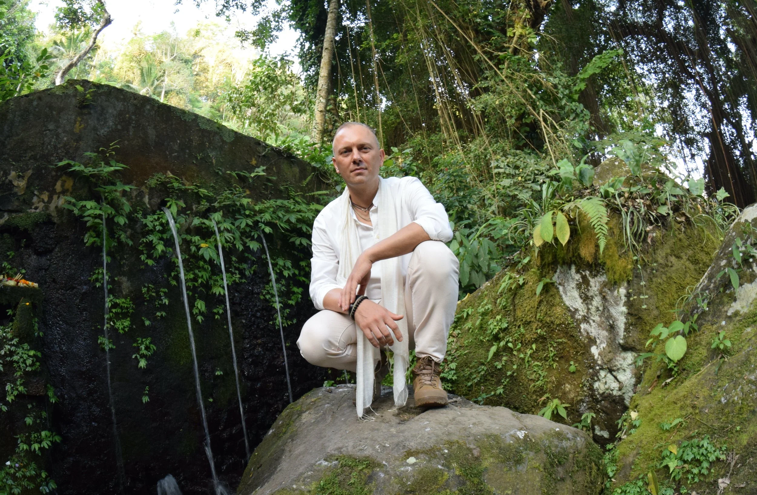 Satthuvi by the river in a lush Balinese jungle, calm and approachable, tropical foliage and flowing water behind; text overlay "Why work with Satthuvi — Purchase Ekinetica Metamorphosis Intensive package."