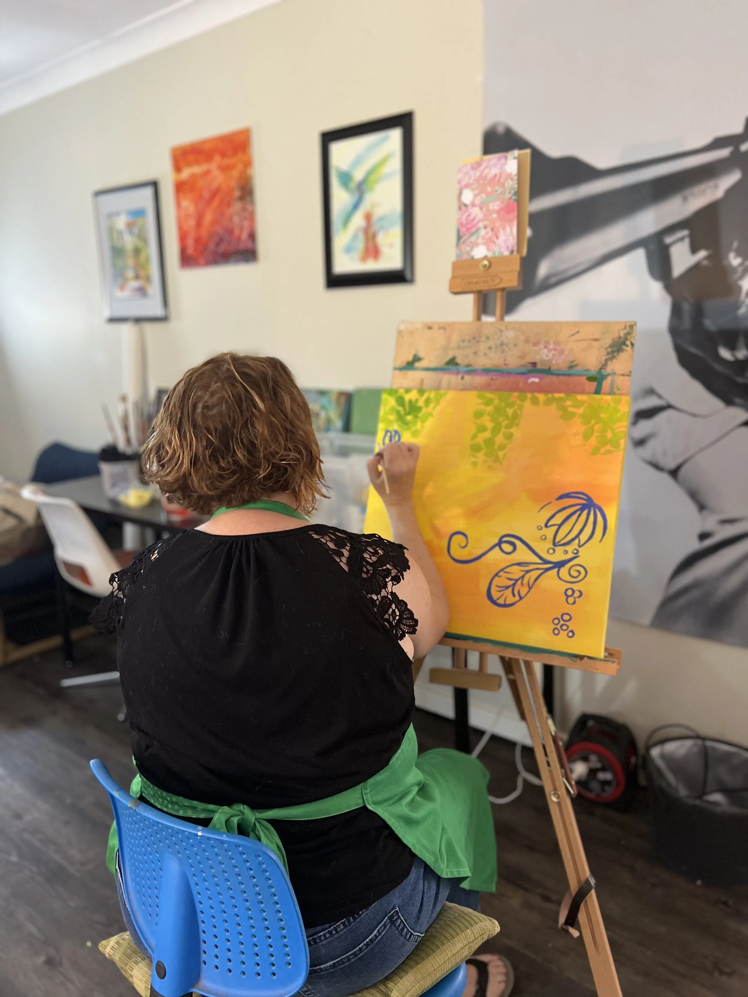 Becca De Jong Art Teacher painting a watercolour canvas. Canvas is yellow with green and blue paint. Becca is seen from behind at the easel in her art studio