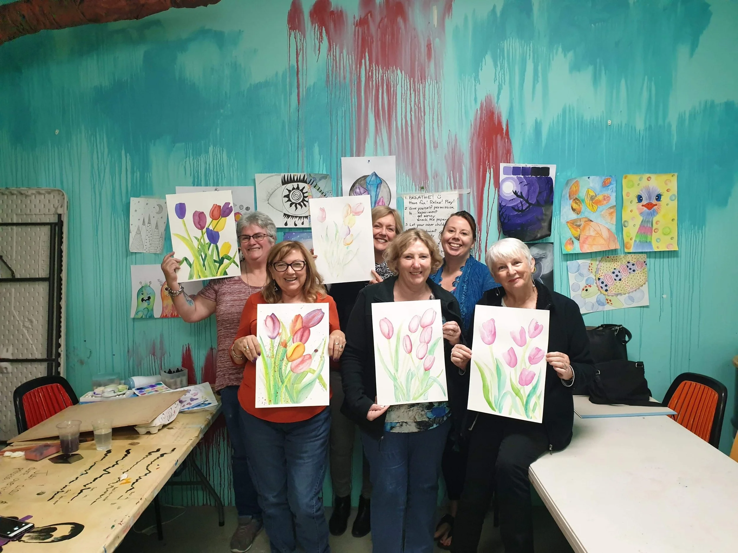 Six women standing in a room with colorful wall art, holding paintings of tulips and other floral artworks, smiling at the camera.