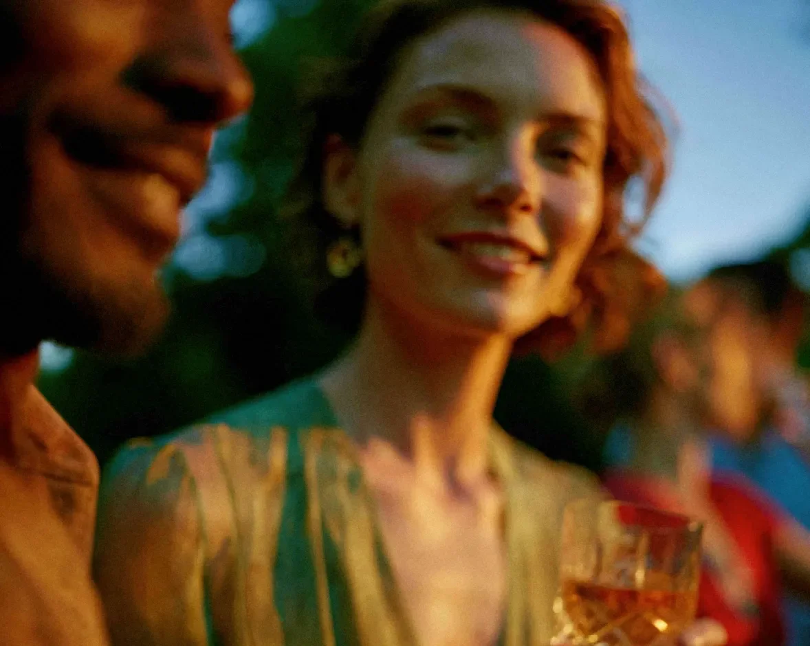 A woman with red hair holding a glass of wine, smiling, with other people at a social gathering in the background.