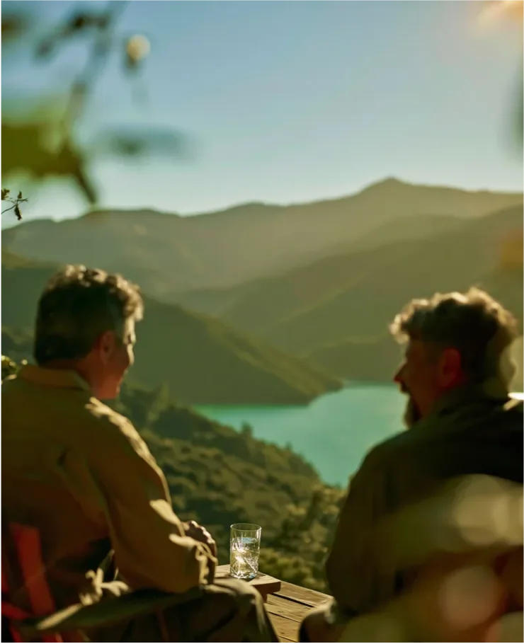 Two men sit outdoors on a wooden deck overlooking the Marlborough Sounds enjoying a glass of Arakiko.