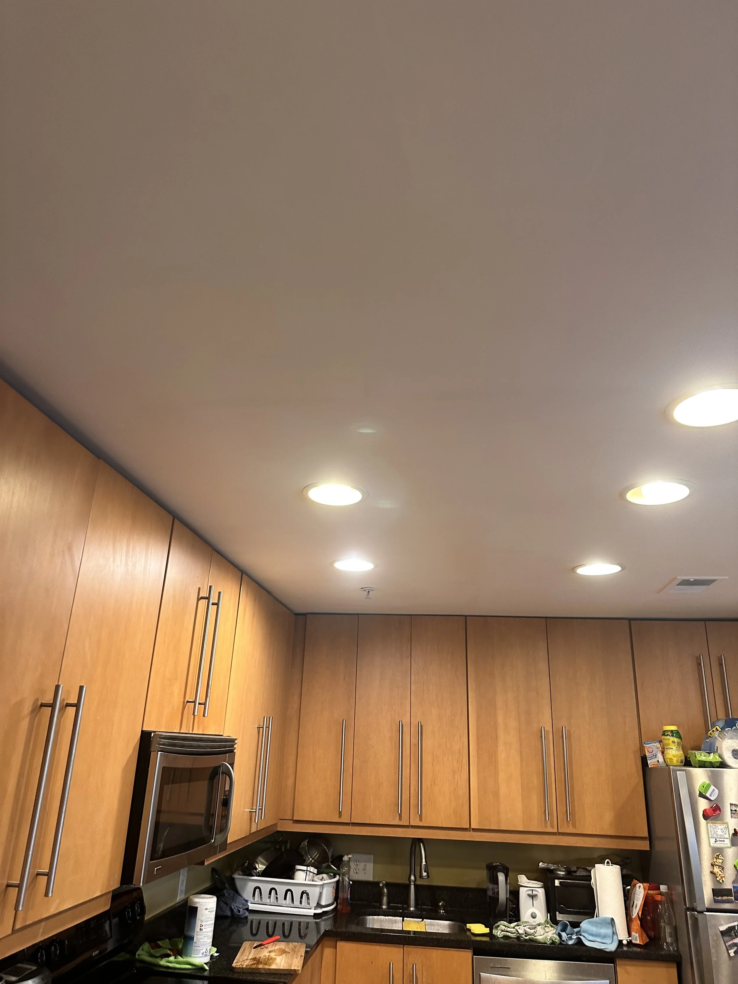 Kitchen with wooden cabinets, stainless steel microwave, refrigerator, various kitchen items, and a tiled countertop.