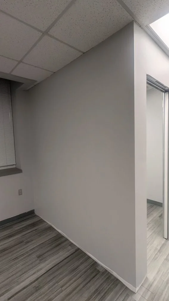 Empty corner of an office with white walls, a window with blinds, and gray wood-patterned carpet.