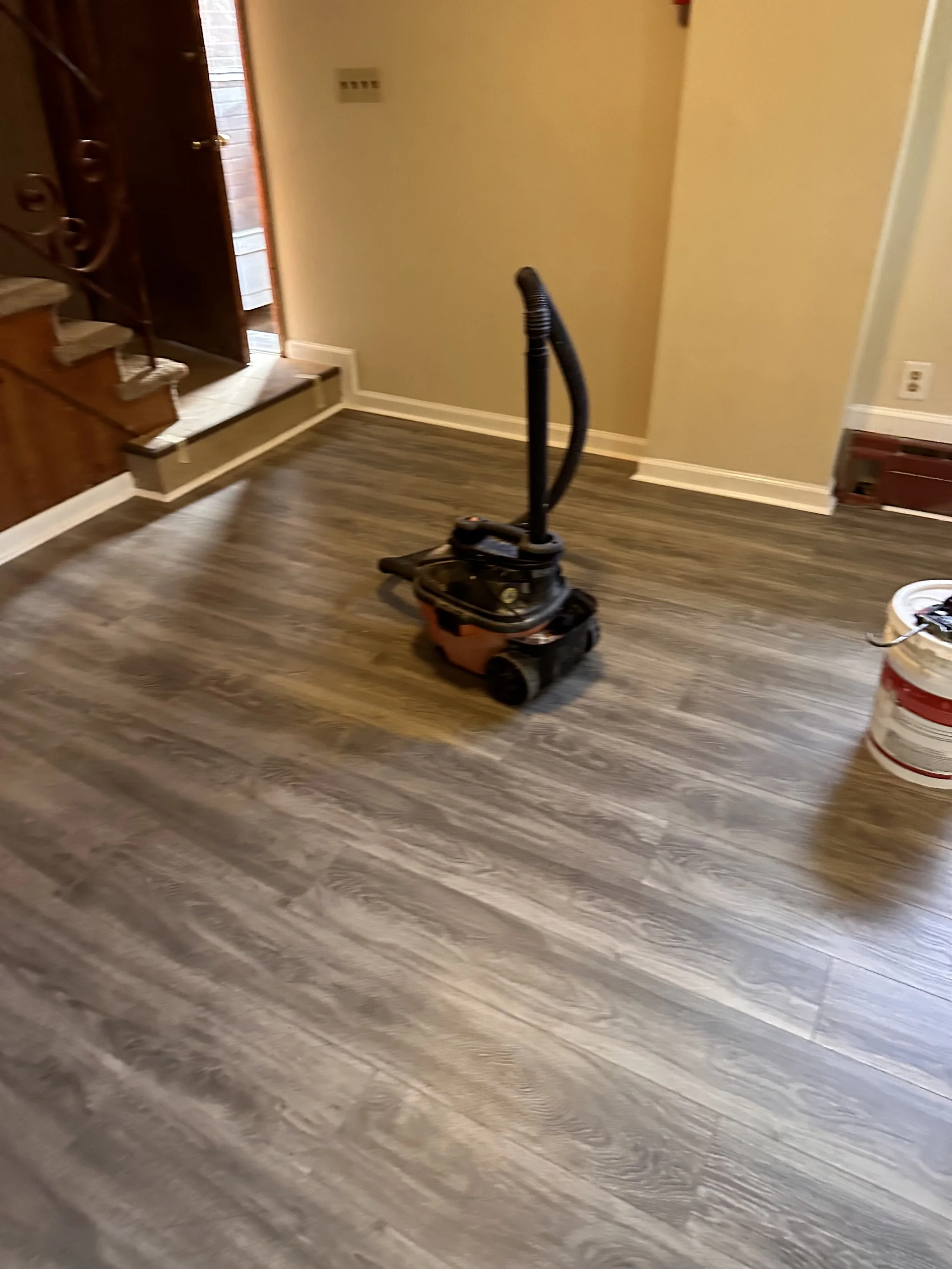 A room with newly installed wood-look vinyl flooring, a vacuum cleaner in the center, and a bucket on the right side.