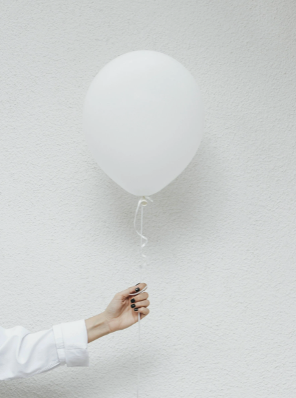 Minimal image of a hand holding a white balloon against a textured wall — symbolising lightness, order, and creative balance.