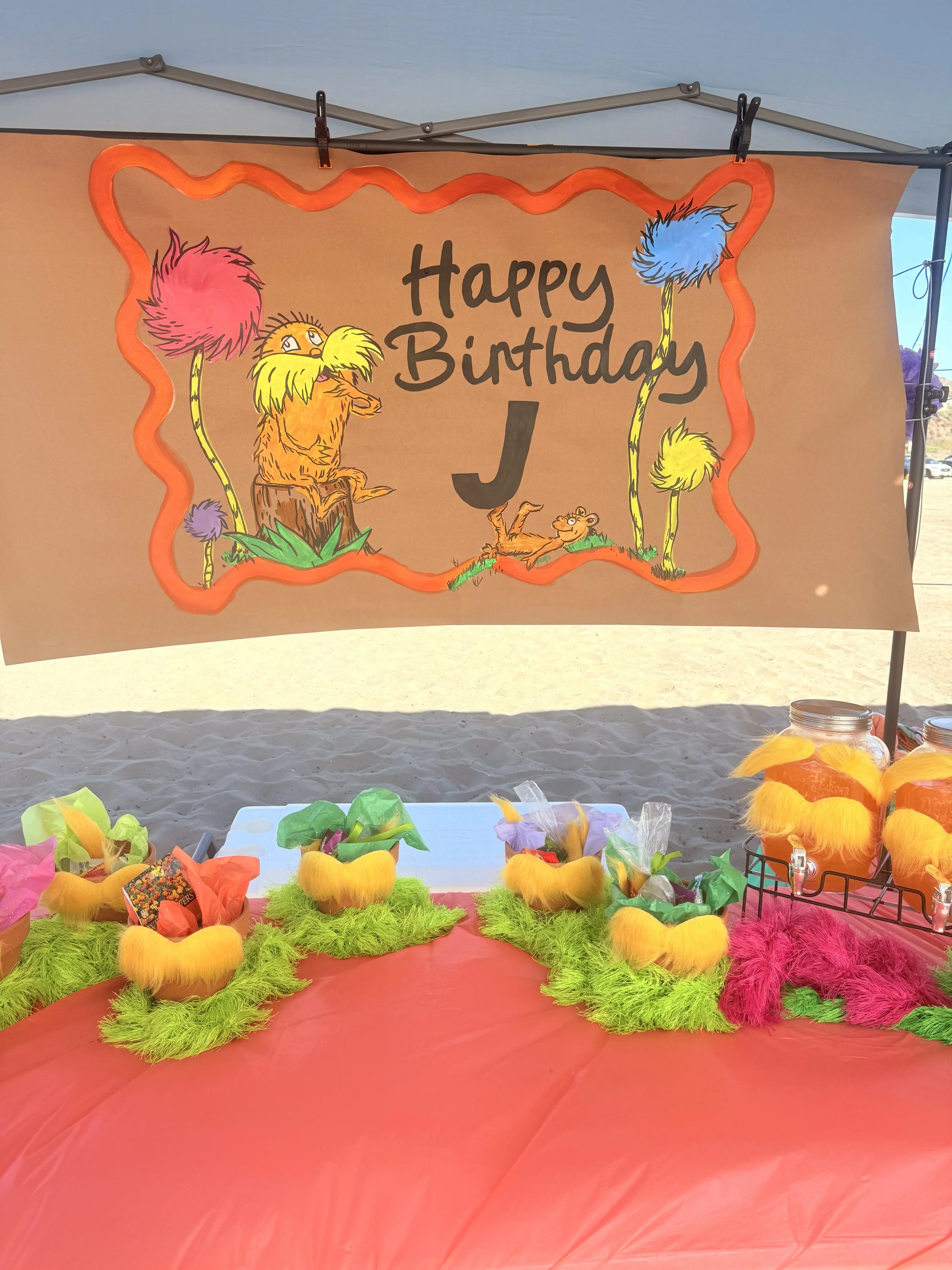 Colorful birthday banner with 'Happy Birthday J' and illustration of the Lorax and Truffula trees from Dr. Seuss, displayed at a beachside party with decorated treat containers.