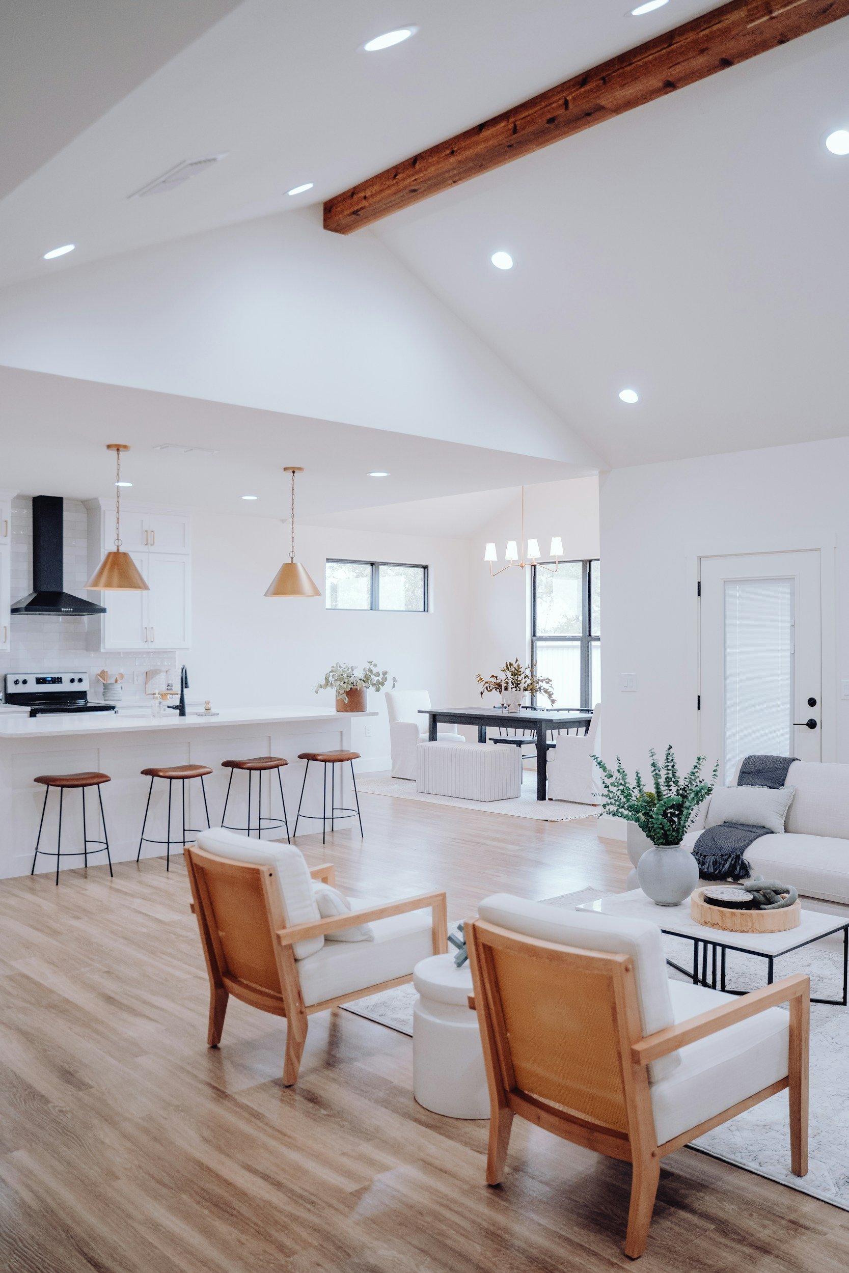 Bright, modern living room and kitchen with white walls, wooden flooring, and minimalist furniture, featuring a seating area with two wooden armchairs and a white sofa, a black coffee table, a dining table with white chairs, and a kitchen island with barstools.