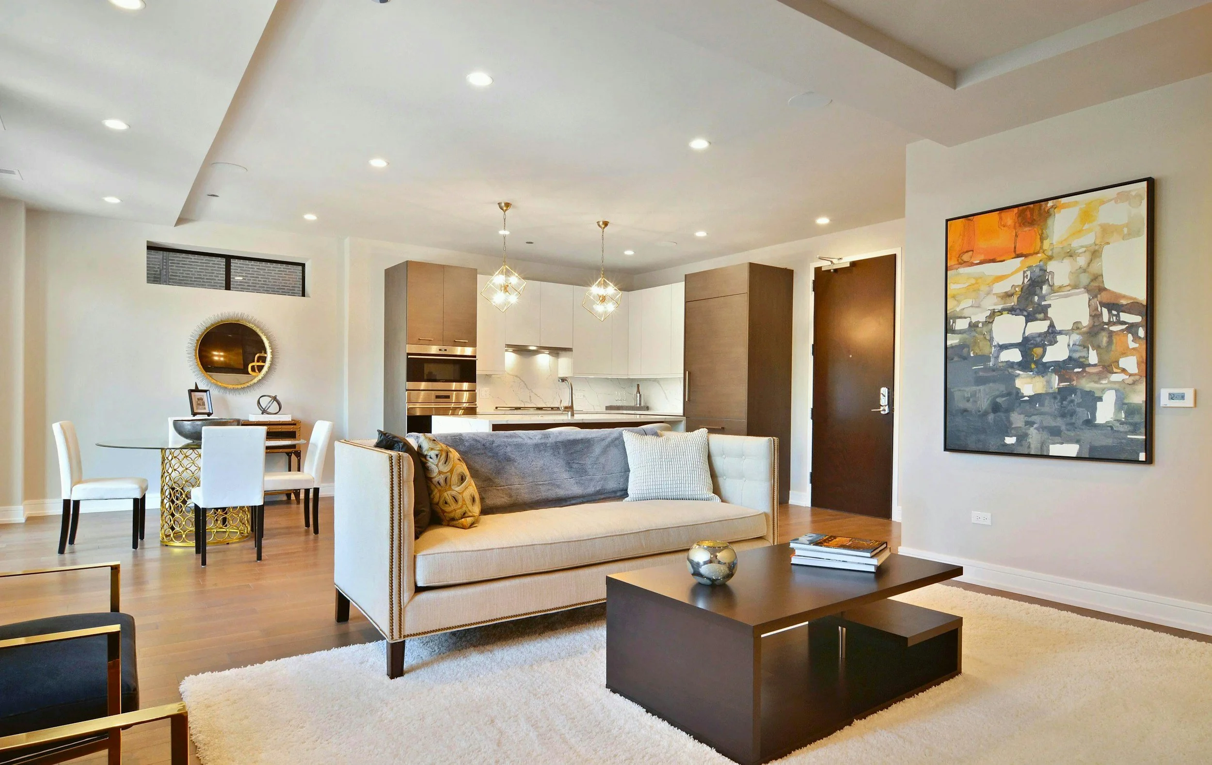 Modern open-concept living room and kitchen with white walls, hardwood floors, beige sofa with pillows, dark wood coffee table, colorful abstract painting, dining area with white chairs, pendant lights, and kitchen with white cabinets, marble backsplash, and stainless steel appliances.