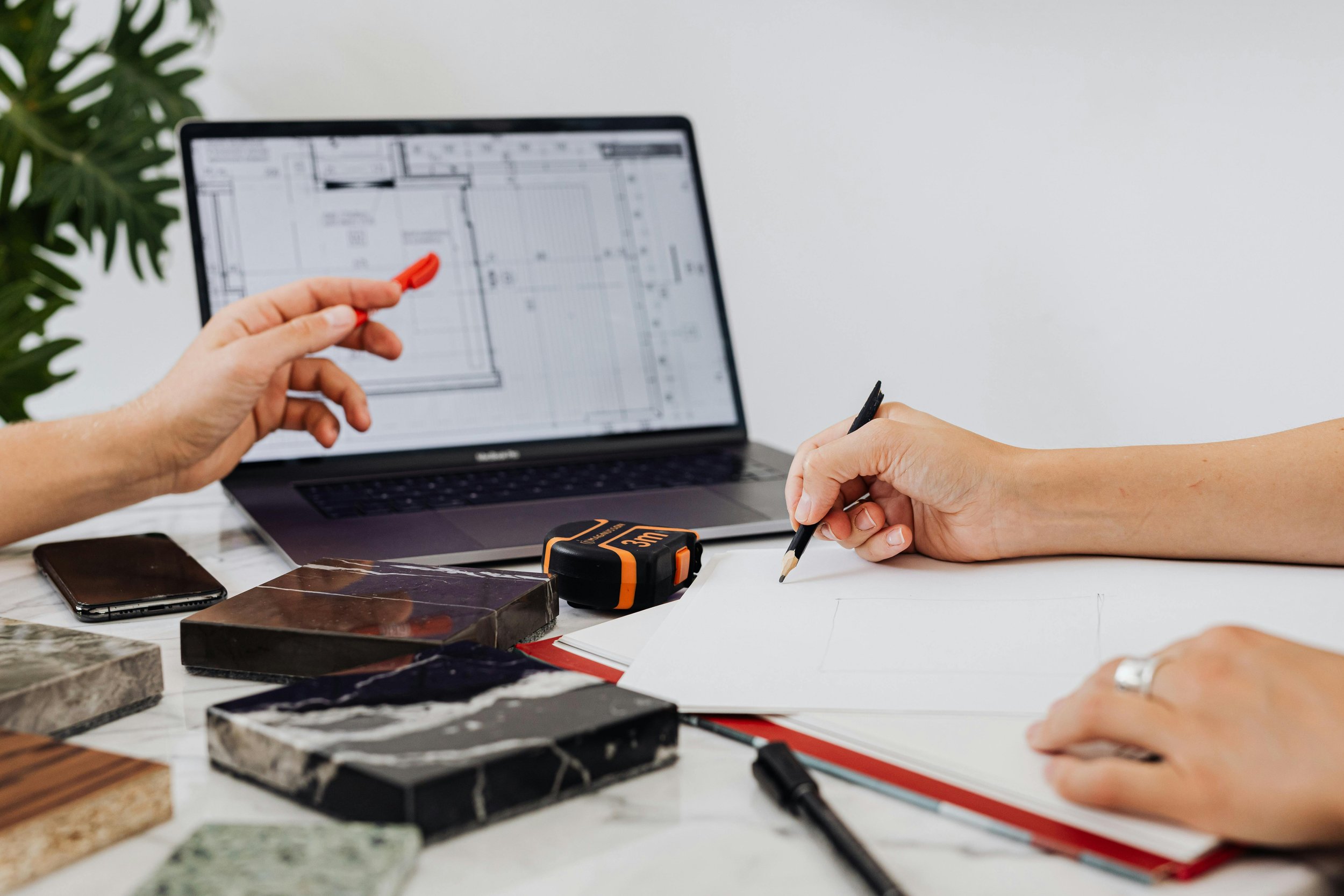 Two people working on a house design at a table with a laptop displaying a blueprint, measuring tape, color samples, and notebooks.