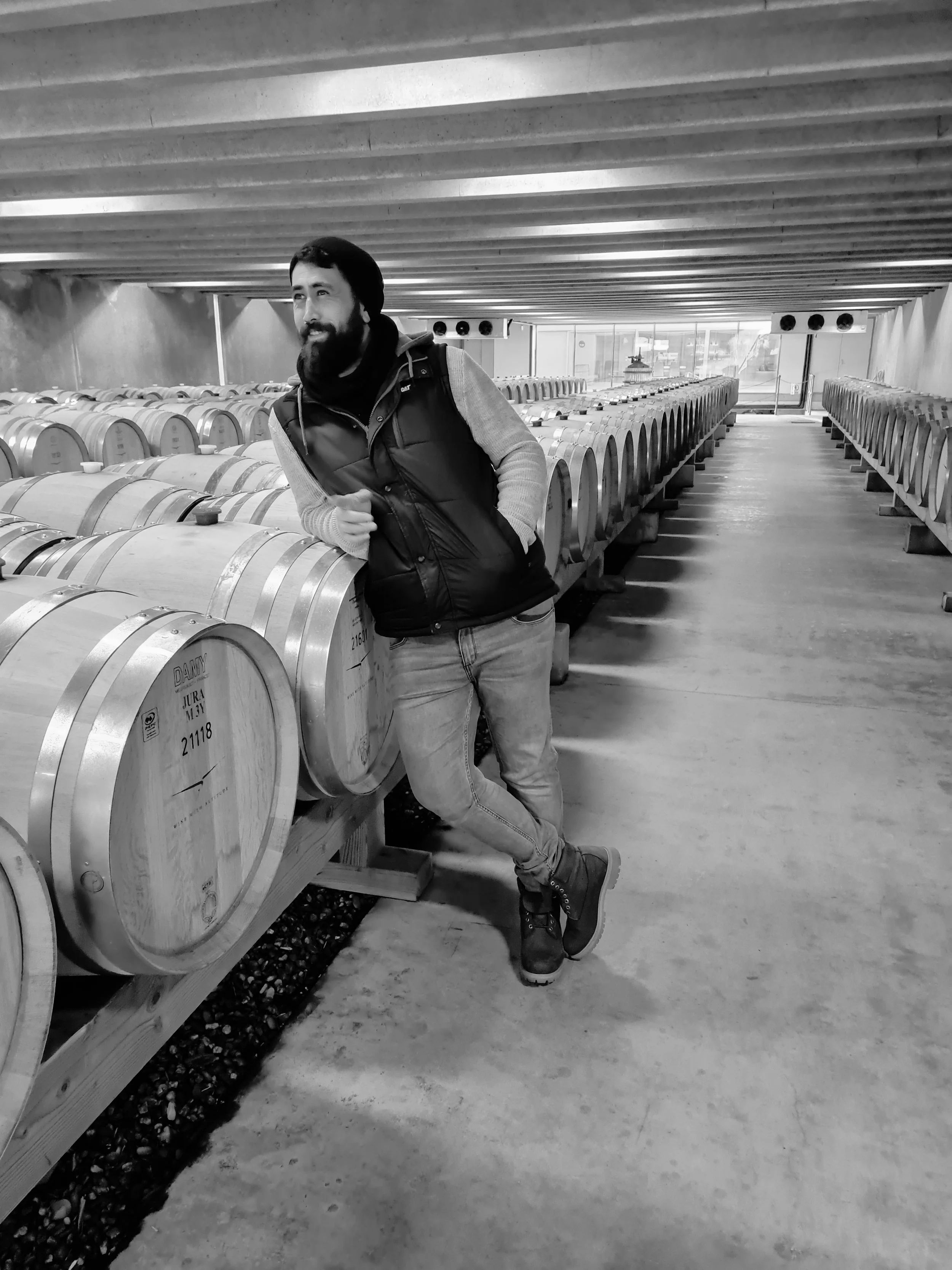 A man with a beard standing inside a winery, leaning on wooden wine barrels, wearing a black vest, gray sweater, jeans, and brown boots.