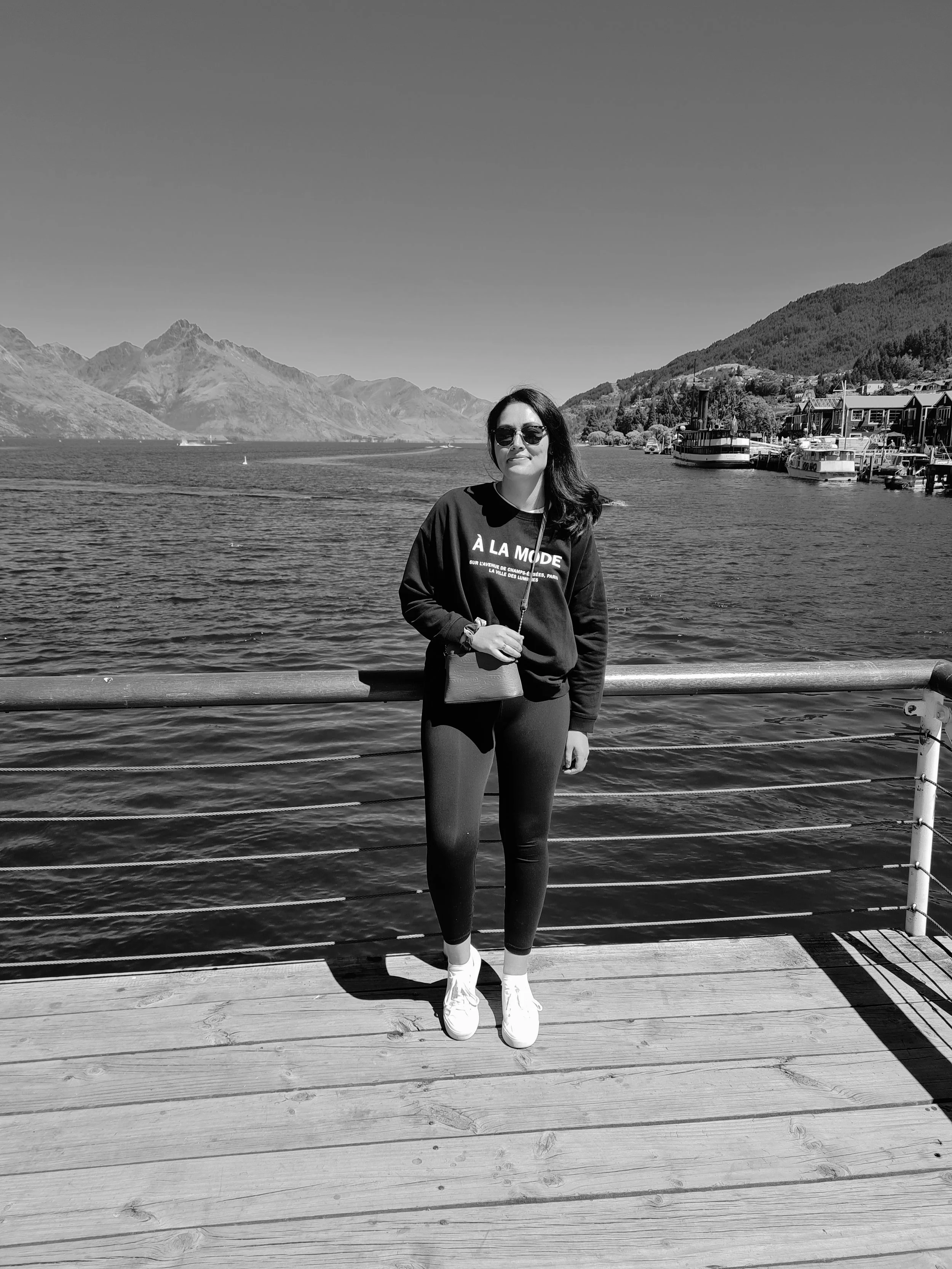 A woman in black leggings, a black sweatshirt, and white sneakers standing on a wooden dock with a scenic lake, mountains, and boats in the background.