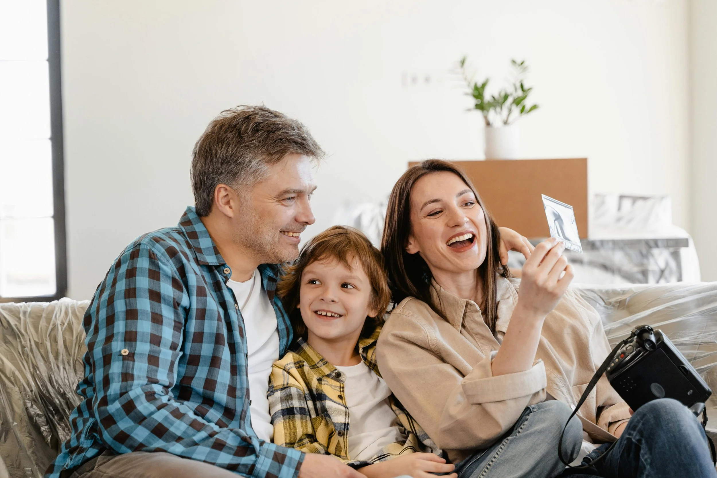 A family of three, a father, mother, and young son, sitting on a couch in a bright living room, smiling and looking at a photograph the mother is holding, with the father and son also smiling.