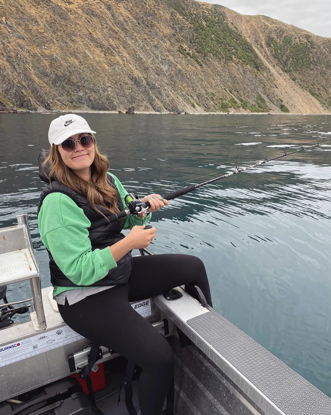 A woman in black leggings, a black sweatshirt, and white sneakers standing on a wooden dock with a scenic lake, mountains, and boats in the background.