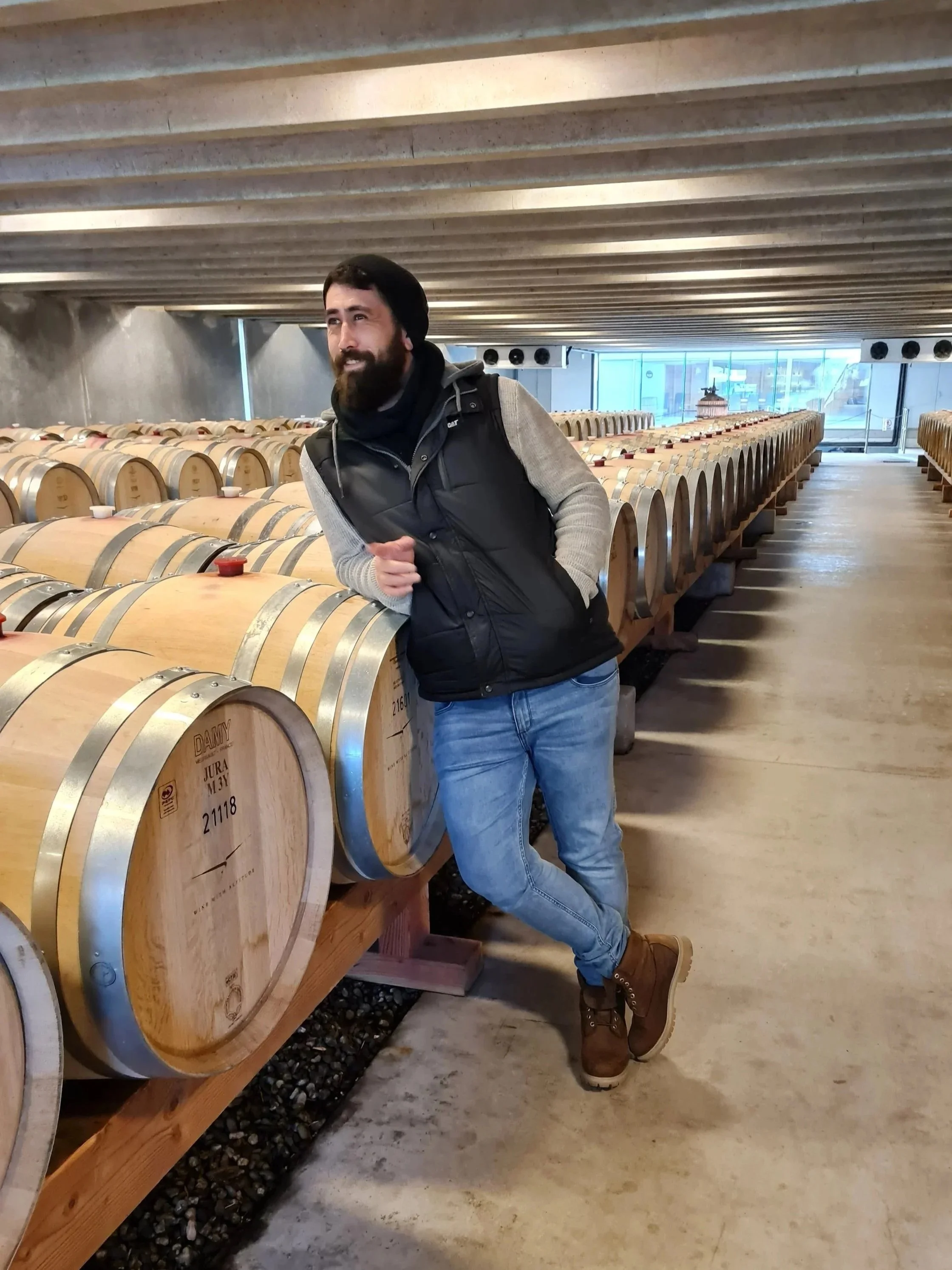 A man with a beard standing inside a winery, leaning on wooden wine barrels, wearing a black vest, gray sweater, jeans, and brown boots.