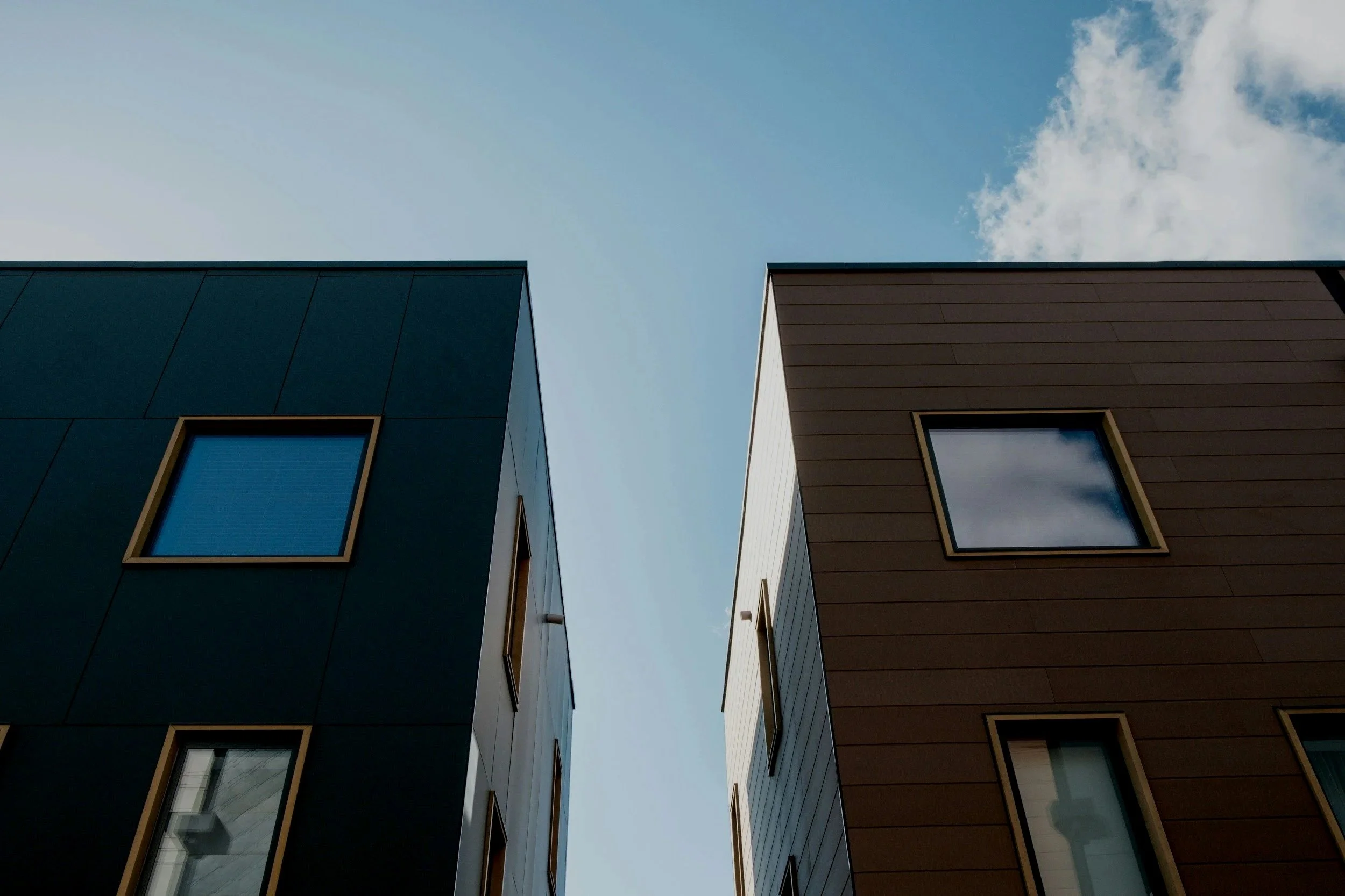 Two modern buildings with dark and brown exteriors and rectangular windows, under a partly cloudy sky.