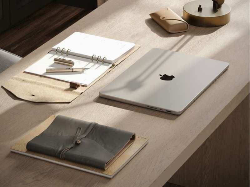 A wooden desk with an Apple MacBook, a closed leather-bound notebook, an open planner with a pen, a tan pencil case, a decorative tray, and a small round clock with a stand.