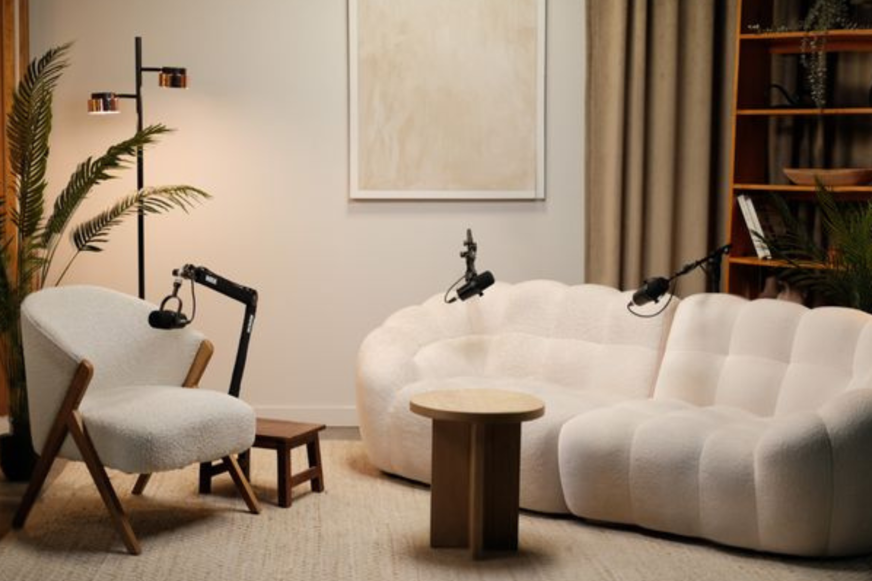 Living room with a cream-colored sofa, white armchair with wooden legs, a small round wooden table, a floor lamp, artwork on the wall, and a bookshelf with plants and decorations.