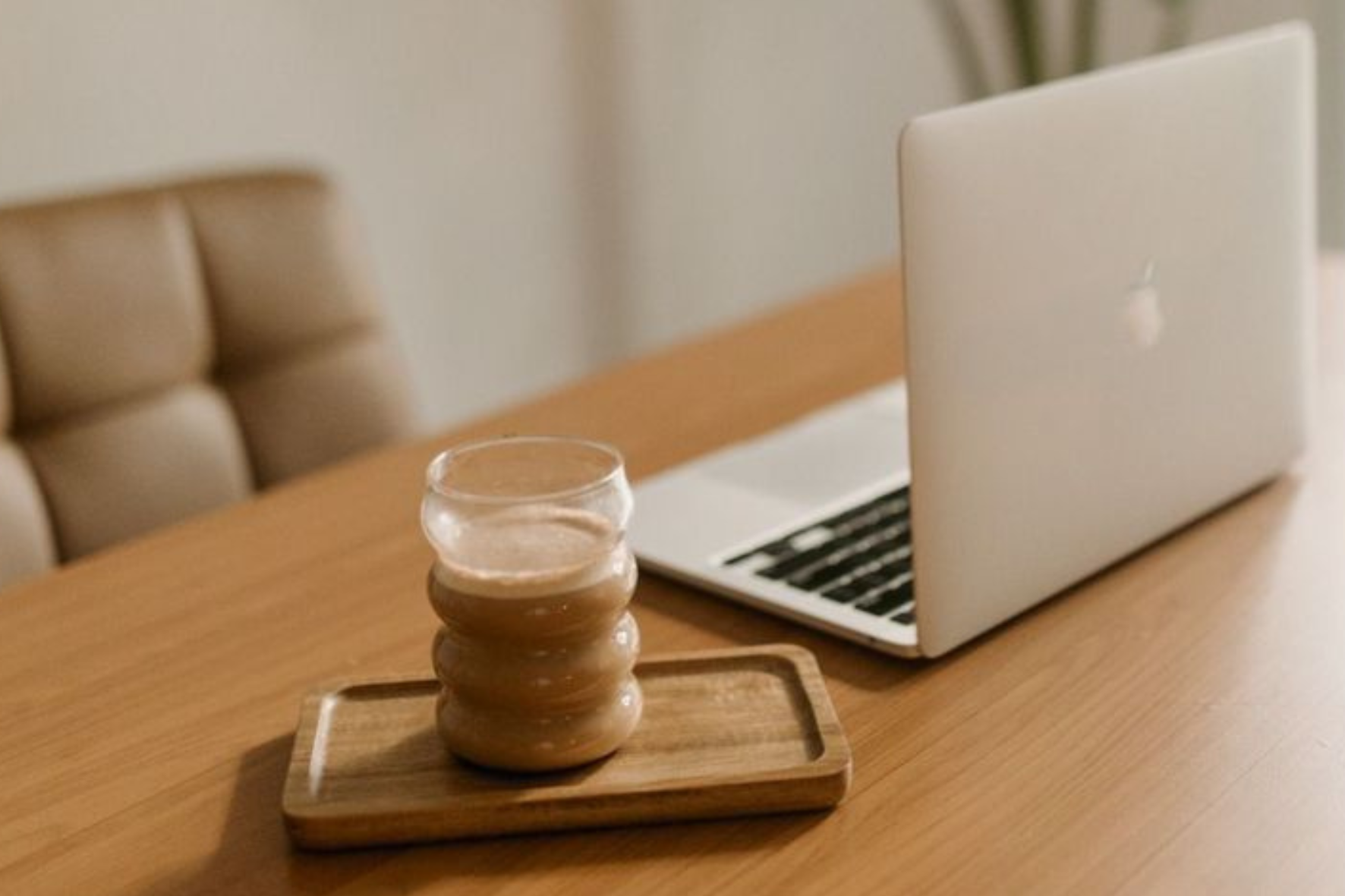 A laptop and a glass of water on a wooden table near a beige cushioned chair.