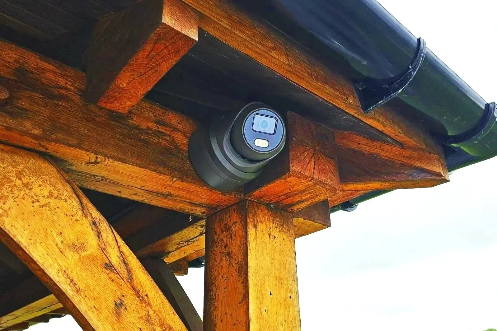 A black wildlife camera mounted under a wooden structure, with wooden beams and a dark green metal roof above it.